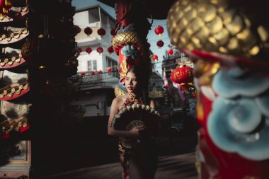 Çin tapınağında geleneksel elbise giymiş Asyalı sakin bir kadın yelpaze tutuyor. Kültür festivali sahnesinde altın ejderha sütunu ve kırmızı fener süslemesi festival havası yaratıyor.