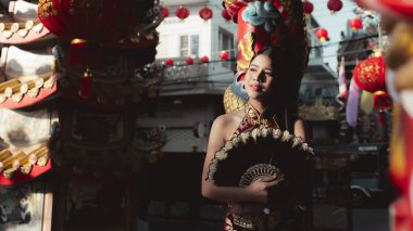Renkli festival kostümü giymiş güzel Asyalı kadın gece canlı pazarda süslü bir hayran tutuyor. Çin dekorasyonu ve fenerlerle çevrili sakin bir ifadesi var.