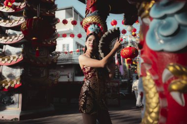 Geleneksel Qipao kıyafetli zarif Asyalı kadın Çin 'in yeni yılını neşeyle kutluyor. Kırmızı fener ve ejderha süslemelerinin ortasında yelpazeyle poz veriyor.