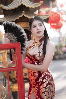 Geleneksel kırmızı Qipao elbiseli zarif Asyalı kadın Çin kültürünün kutlamaları sırasında hayranlarını tutuyor. Düşünceli ve sakin bir ifadesi var. Kameradan başka yere bakıyor.