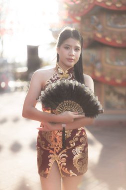 Siyah yelpaze tutan geleneksel kırmızı Çinli elbiseli güzel bir Asyalı kadın. Kültür festivali sırasında sıcak bir ışıkla dışarıda dikilen sakin bir ifadesi var.