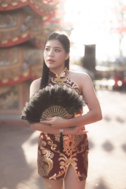 Geleneksel kırmızı ve altın rengi Cheongsam elbiseli güzel Çinli kadın dışarıda yelpaze tutuyor. Kültür ortamında dururken düşünceli bir yüz ifadesi var.