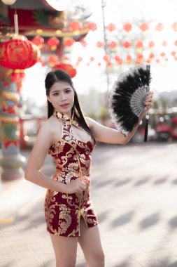 Geleneksel kırmızı Cheongsam elbisesi içinde genç ve güzel bir Asyalı kadın, Çin Yeni Yıl Festivali 'nde fenerlerle mutlu ve kendinden emin bir şekilde siyah yelpaze tutuyor.