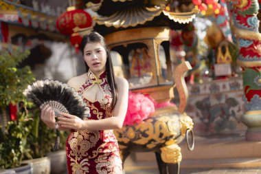 Geleneksel kırmızı Çin elbisesi giymiş sakin genç Asyalı kadın ya da Cheongsam yelpaze tutuyor. Kültür festivali sırasında tapınakta zarafetle poz veriyor.