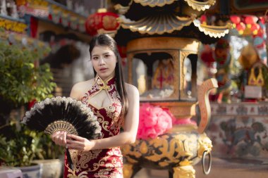 Geleneksel kırmızı Cheongsam elbisesi giymiş, elinde siyah yelpaze tutan sakin Asyalı kadın. Çin tapınağında duruyor. Kültürü ve geleneği sakin ve zarif bir ifadeyle somutlaştırıyor.