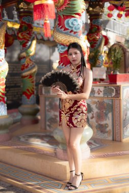 Geleneksel kırmızı Cheongsam elbisesi içindeki kendinden emin genç Asyalı kadın kutlama için birçok fenerle süslenmiş canlı Çin tapınağının içinde siyah yelpaze tutuyor.