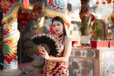 Geleneksel kırmızı altından Cheongsam yelpaze tutan sakin, genç Asyalı kadın. Çin tapınağında güven içinde poz vermek, yeni yıl festivalinde kültürü kutlamak.