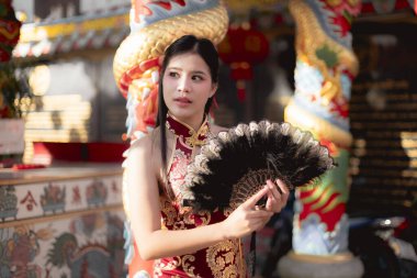 Geleneksel kırmızı Cheongsam elbiseli güzel Asyalı kadın Çin tapınağında yelpaze tutuyor. Onun sakin ifadesi kültürel zarafeti ve geleneği yansıtıyor.