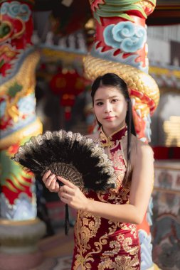 Geleneksel kırmızı Qipao elbise giymiş güzel bir Çinli kadın elinde yelpaze tutuyor. Renkli bir tapınakta duran huzurlu bir yüzü olan zarif bir portresi.