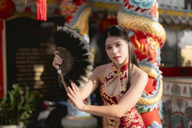 Geleneksel kırmızı Cheongsam 'lı güzel Asyalı kadın elinde siyah yelpaze tutuyor. Çin kültürünü ve yeni yıl festivalini kutlamak için ejderha heykeliyle poz veren zarif kız.