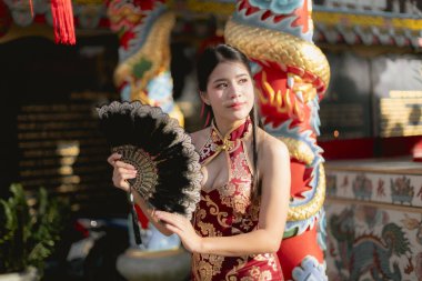 Geleneksel Çin Qipao elbisesi giymiş güzel Asyalı kadın tapınakta yelpaze tutuyor. Yüzünde zarif ve zarif bir ifadeyle kültürün huzurlu bir kutlaması.