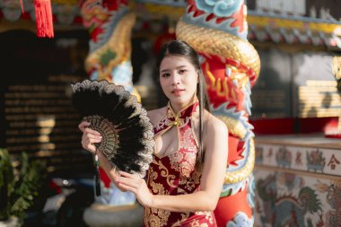 Siyah yelpaze tutan kırmızı geleneksel Cheongsam elbiseli kendinden emin genç Asyalı kadın. Kültür festivali sırasında Çin tapınağında poz veren güzel bir model.