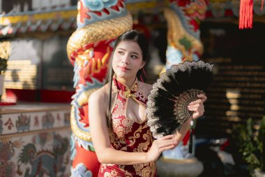 Geleneksel kırmızı Cheongsam elbiseli güzel Çinli kadın elinde siyah yelpaze, zarif bir şekilde tapınakta poz veriyor. Bu sahne Asya kültürünü ve kutlamalarını temsil ediyor.