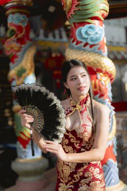 Geleneksel kırmızı Qipao elbisesi içindeki zarif Çinli kadın tapınakta siyah yelpaze tutuyor ve yüzünde zarif bir ifadeyle Asya kültürünü kutluyor.