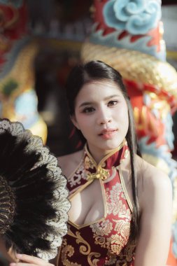 Kırmızı Cheongsam elbiseli sakin Asyalı kadın tapınakta renkli Çin ejderhası heykeliyle poz verirken siyah yelpaze tutuyor, kameraya güvenle bakıyor.