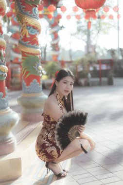 Geleneksel Qipao 'lu zarif Asyalı kadın renkli Çin tapınağında çömeliyor. Bu portre onun yelpazesini sakin ve cezbedici bir ifadeyle çeker.