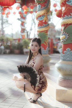 Geleneksel Cheongsam elbisesi içindeki mutlu Asyalı kadın tapınakta zarif bir şekilde gülümseyen siyah yelpaze tutuyor. Şenlikli kırmızı fenerler dışarıdaki kültürel kutlamaları süslüyor.