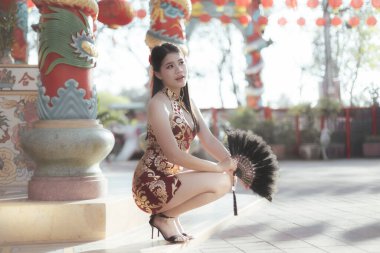 Geleneksel kırmızı Cheongsam elbiseli zarif Asyalı kadın, Çin tapınağında hayranlarıyla poz veriyor, festival sırasında kültürel zarafet ve sükuneti somutlaştırıyor.