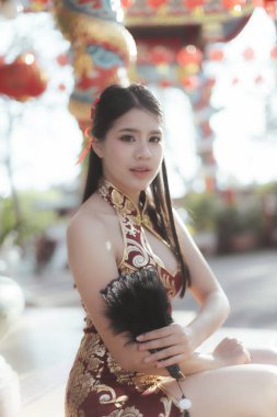 Geleneksel kırmızı Cheongsam elbiseli sakin genç Asyalı kadın Çin Yeni Yıl kutlamaları sırasında açık hava tapınağında tüy hayranıyla zarif poz veriyor.