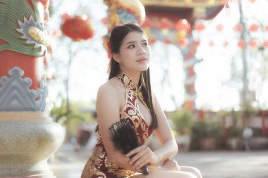 Güzel geleneksel Çin Qipao elbisesi içindeki düşünceli, dingin genç Asyalı kadın arka planda kırmızı fenerle süslenmiş tapınakta otururken yelpaze tutuyor.
