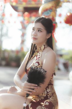 Geleneksel kırmızı Çin Qipao elbisesi içinde güzel bir Asyalı kadın tapınakta yeni yıl kutlaması sırasında huzurlu görünüyor. Elinde festival fenerleriyle çevrili siyah bir yelpaze var.