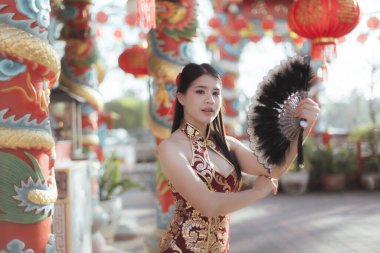 Geleneksel kırmızı Qipao giyen zarif genç Asyalı kadın Çin Yeni Yıl kutlamaları için elinde siyah yelpaze tutuyor. Tapınakta güzel fenerle süslenmiş.