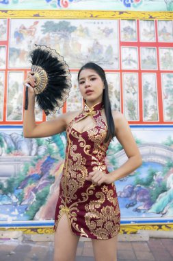 Geleneksel kırmızı Cheongsam Qipao elbisesi içindeki güzel Asyalı kadın kendinden emin poz veriyor. Renkli Çin duvar resmine karşı siyah yelpaze tutuyor. Zarif görünüyor.