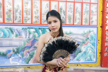 Kırmızı geleneksel elbiseli güzel Asyalı kadın siyah el yelpazesiyle zarif poz veriyor, renkli tapınağın içinde kendinden emin bir ifadeyle kameraya bakıyor.