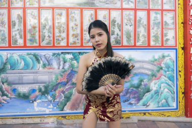 Geleneksel kırmızı Cheongsam elbisesi içindeki güzel Asyalı kadın siyah yelpaze tutuyor, renkli Çin tapınağında antik duvar resimleriyle güvenle poz veriyor.