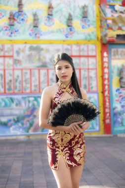 Siyah el yelpazeli modern kırmızı Qipao elbiseli zarif Asyalı kadın kendine güvenerek poz veriyor. Canlı, renkli bir tapınak duvarının önünde duruyor. Geleneksel sanatla dolu.