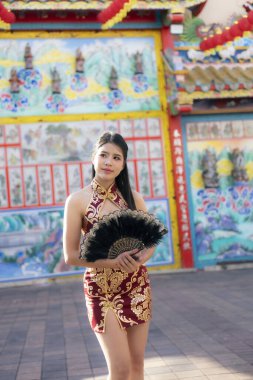 Geleneksel kırmızı Qipao elbise giymiş güzel bir Asyalı kadın elinde yelpaze tutuyor. Dışarıdaki renkli, süslü Çin tapınağının önünde kendine güvenerek duruyor.