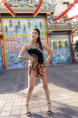 Modern geleneksel Cheongsam elbiseli güzel Asyalı kadın kendine güvenen bir yelpaze ile poz veriyor. Kültürel görünüşü renkli Çin tapınağının önünde yankılanıyor.