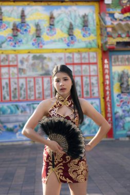 Kırmızı geleneksel elbiseli kendine güvenen Asyalı kadın yelpaze tutuyor ve cazibeyle poz veriyor. Güzel manken renkli kültürel arka plana karşı açık havada duruyor.