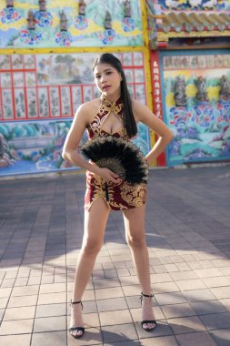 Modern kırmızı Qipao elbiseli kendine güvenen genç Asyalı kadın dışarıda poz veriyor. Seçkin kadın modelin elinde siyah yelpaze var. Şehir sokağında duruyor. Geleneksel moda sergiliyor.