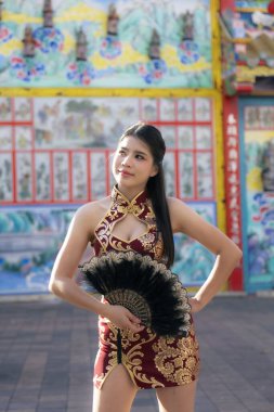 Geleneksel kırmızı Cheongsam elbisesi içinde siyah yelpaze tutan kendine güvenen güzel Asyalı kadın. Çin tapınağında sakin bir ifadeyle poz veren zarif bir kadın model.