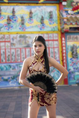 Modern kırmızı ve altın rengi Cheongsam ya da Qipao 'lu kendine güvenen Asyalı kadın geleneksel hayran kitlesine sahip. Tarzı ve tarzı tapınakta poz verirken kültürünü yansıtıyor.