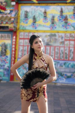 Geleneksel kırmızı elbiseli güzel Asyalı kadın yelpaze tutuyor, düşünceli ifadesi yukarı bakıyor. Renkli tapınağın önünde duruyor, kültürle modayı harmanlıyor.