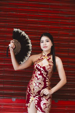 Geleneksel kırmızı Çin cheongsam elbisesi giymiş güzel bir Asyalı kadın siyah yelpaze tutuyor. Neşeli kırmızı arka planda festival kültürünü kutlayan kendine güvenen bir gülümseme