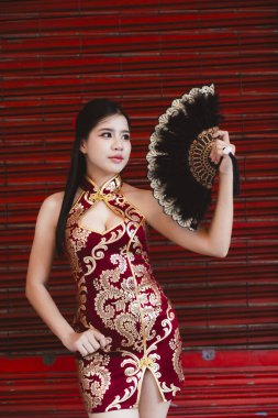 Siyah yelpaze tutan geleneksel kırmızı ve altın rengi Cheongsam Qipao elbiseli zarif Asyalı kadın. Kendine güveni tam, kutlama için kırmızı arka planda duruyor.