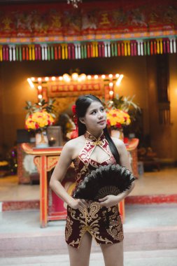 Geleneksel Qipao elbisesi giymiş güzel bir Asyalı kadın elinde vantilatörle tapınağın içinde duruyor. Düşünceli ifadesi kültür festivali kutlamalarında saygı gösterdiğini gösteriyor.