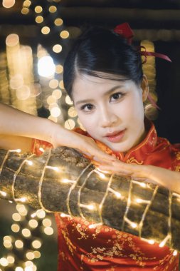 Geleneksel kırmızı elbiseli güzel Asyalı kadın festival gecesi kutlamalarında zarif bir şekilde gülümsüyor. Parlak dekoratif peri ışıklarıyla poz veriyor.