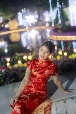 Güzel, genç Asyalı bir kadın geleneksel kırmızı Cheongsam elbisesi giyiyor. Huzurlu bir gülümseme ile gölün kenarında oturuyor. Arka planda kameraya bakıyor.