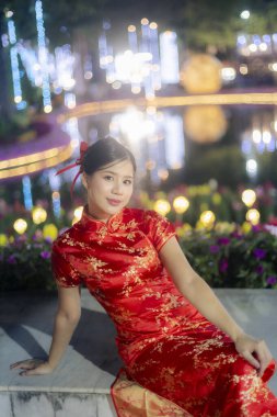Kırmızı Cheongsam elbisesi giyen güzel bir Asyalı kadın kutlama festivali sırasında şık ve renkli arka planda oturuyor.