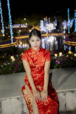 Genç mutlu Asyalı kadın geceleri dışarıda oturuyor. Kırmızı geleneksel Cheongsam elbisesi giyiyor. Arka planda bokeh ışığı var.