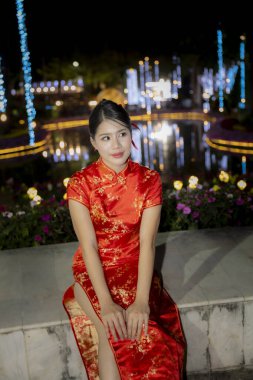 Geleneksel kırmızı Cheongsam elbisesi giyen güzel Asyalı bir kadın kutlama gecesinde yüzünde hafif bir gülümsemeyle dışarıda oturuyor.