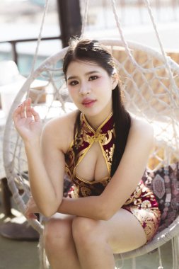 Şık kırmızı Qipao elbiseli, kendine güvenen genç Asyalı kadın, zarif bir görünüşle poz veriyor. Çekici bir ifadeye sahip güzel bir Çin modeli modern sallanan sandalyede oturur.