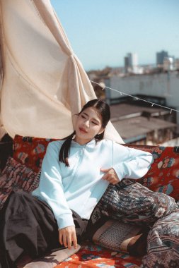 Sakin, genç Asyalı kadın güneşli çatıda dinleniyor. Güzel bir hayat tarzı, sakin bir ifadeyle şehir manzarasından hoşlanan birinin portresi.