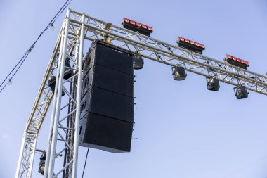 Profesyonel ses sistemi hoparlörleri ve metal kiriş yapısına asılı ışıklarla açık hava konser sahnesi, gösteri için güçlü bir beklenti hissi uyandırıyor