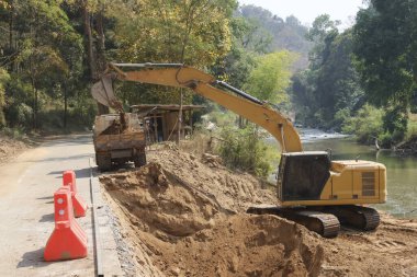 Yol inşaatında ağır makineler çalışıyor. Kırsal alanda altyapı onarımı ve geliştirilmesi için toprak kazıcı sarı kazıcı ve çöp kamyonuna kum yükleme