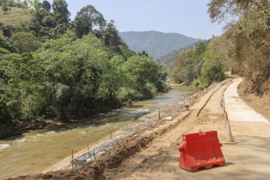 Kırsal dağ manzarasında akan nehrin yanında yeni beton yol inşaatı projesi. Bu sakin manzara uzak doğa alanındaki altyapı gelişimini gösteriyor